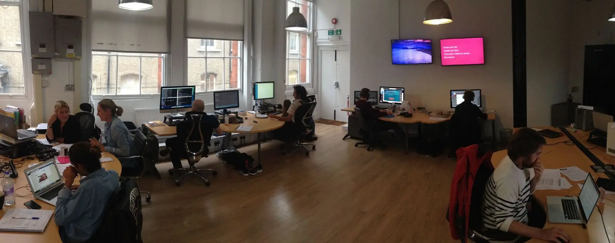 A panorama of the Old Street studio: people at curved desks under pendant lamps, and two ScreenCloud screens on the far wall