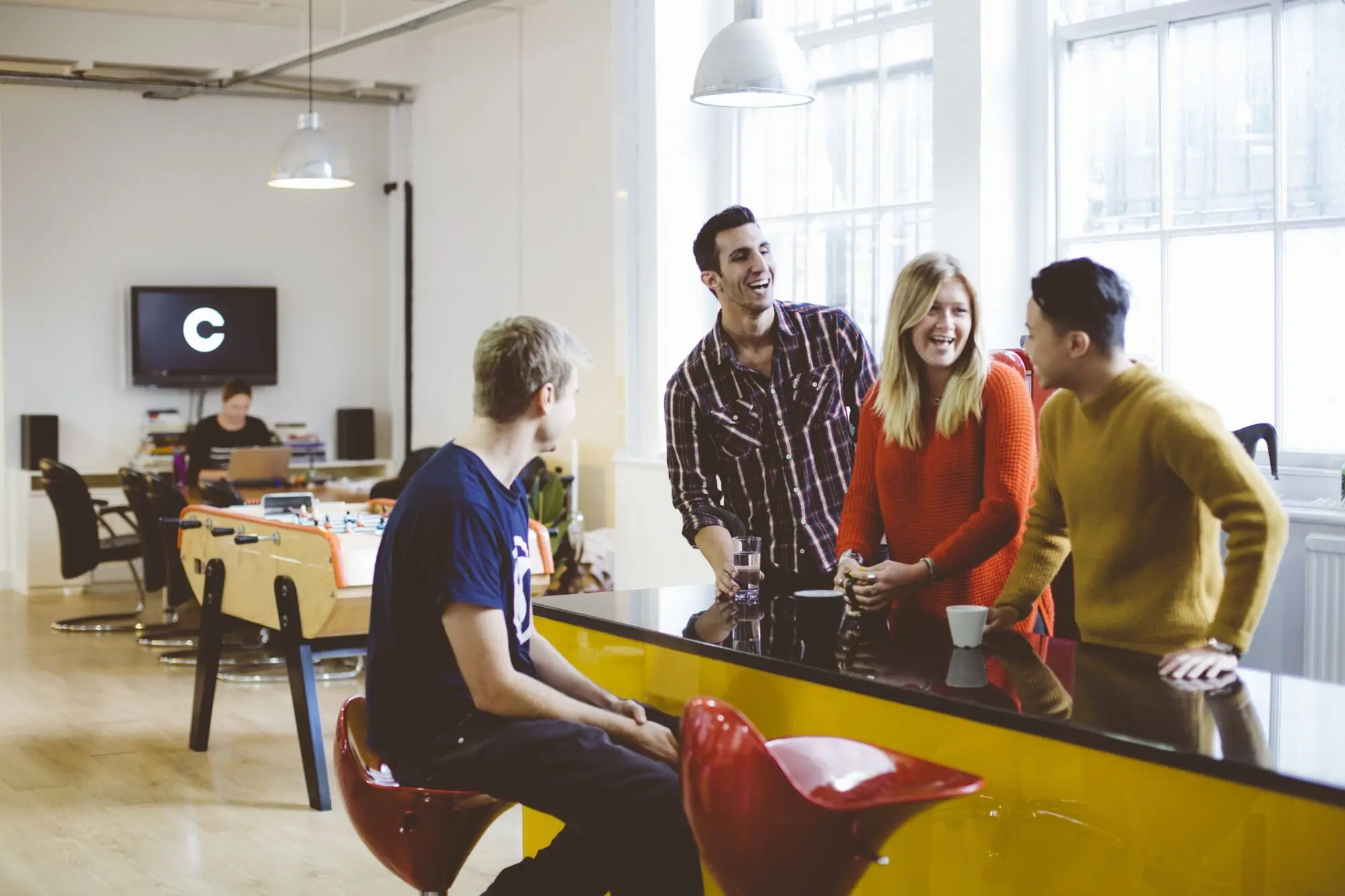 Five of the team laughing at the yellow kitchen counter, with the big C on a screen behind them