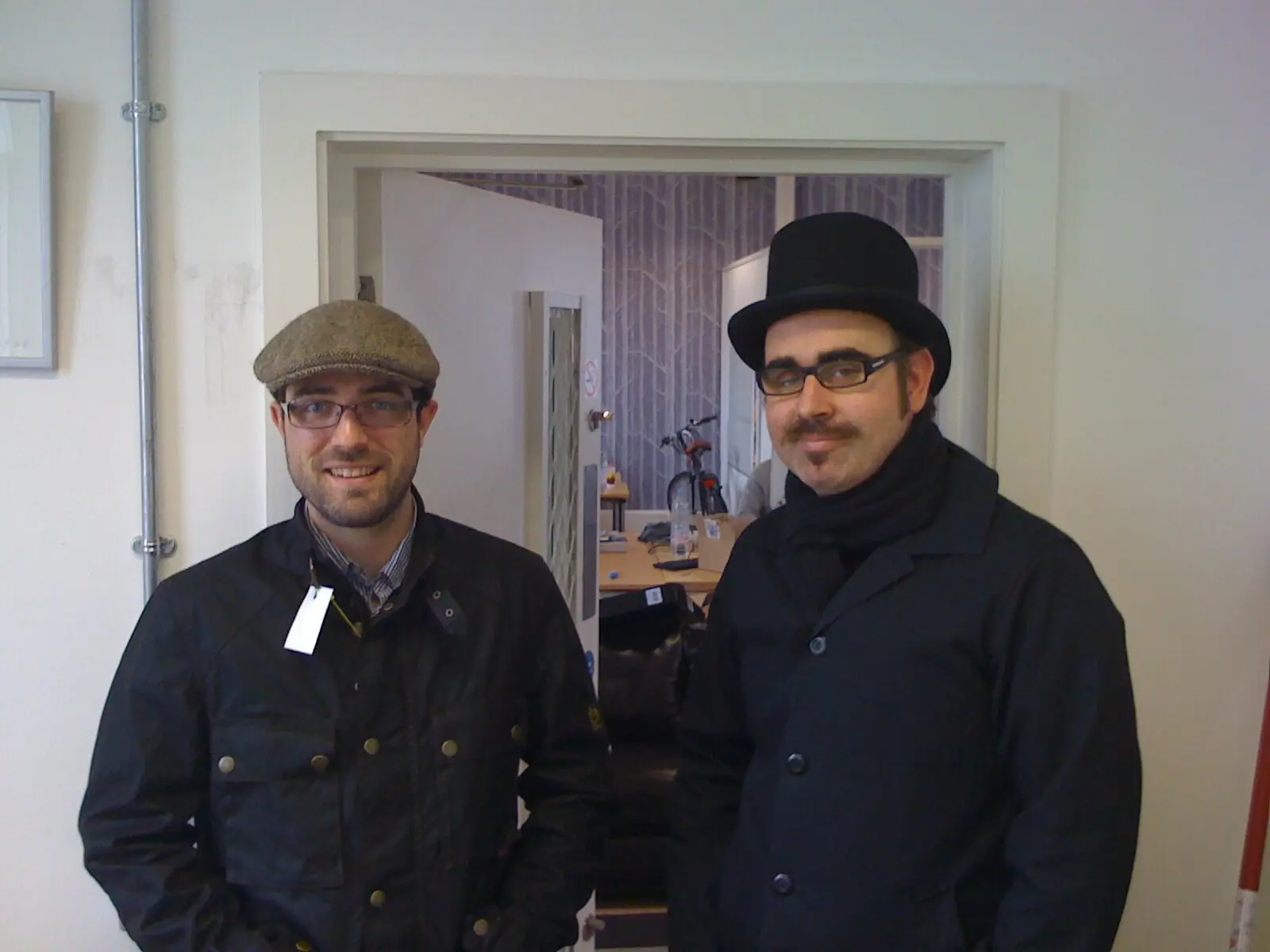 Two of the team in a flat cap and a top hat in a Clapham doorway, the birch-tree wallpaper behind them